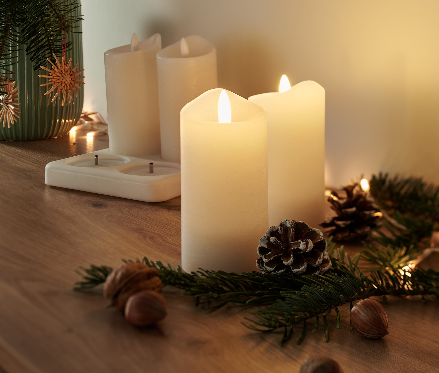 Pilli 4 adet gerçek bal mumu mum, çam dalları, kozalaklar ve fındıklarla dolu bir vazonun yanında ahşap bir masa üzerinde duruyor.