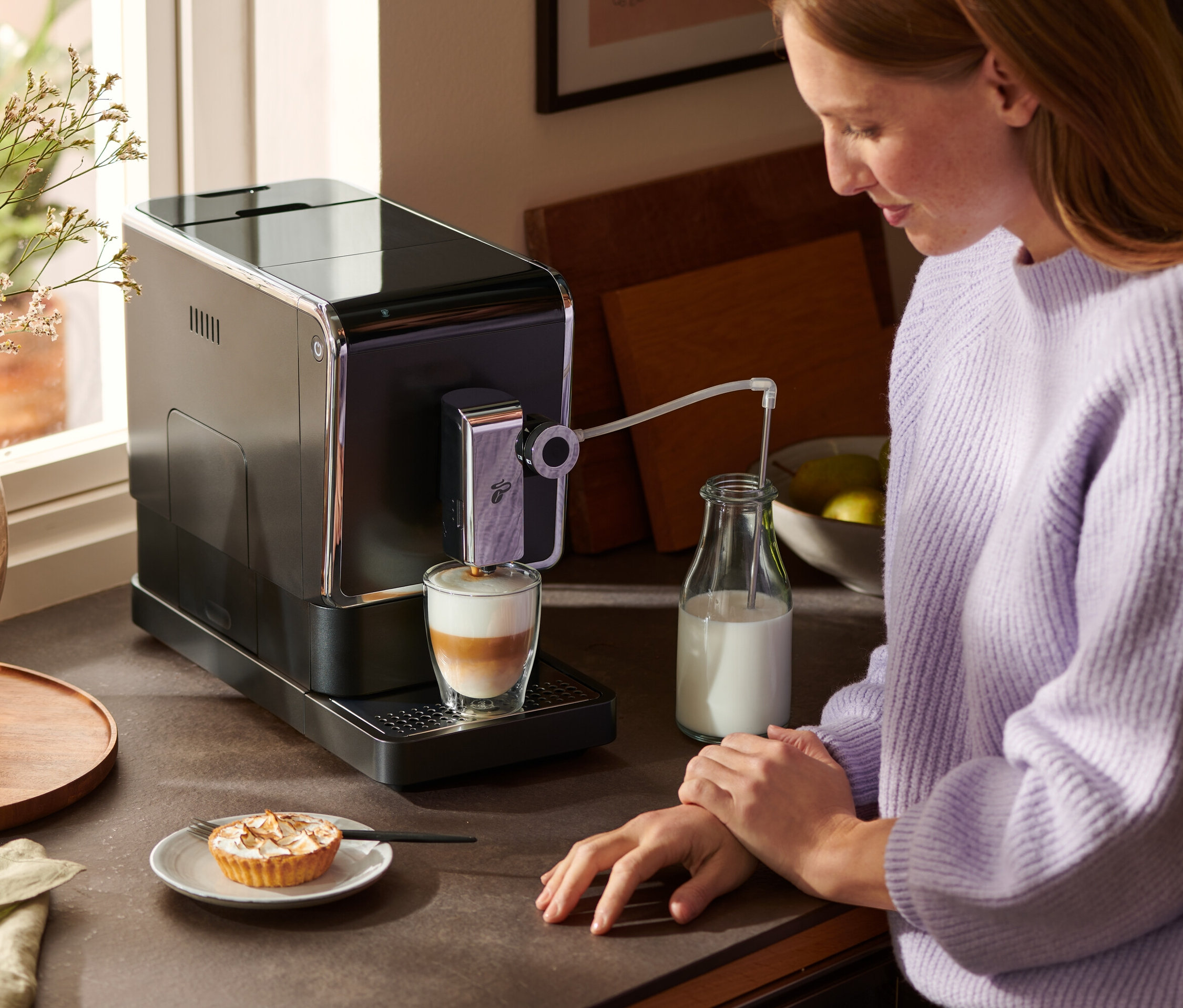 Bir kadın tezgahın yanında duruyor ve antrasit renkli »Esperto Pro« Tchibo tam otomatik kahve makinesi ile latte hazırlıyor. Kahve makinesinin yanında bir süt şişesi ve bir tabak kek duruyor.