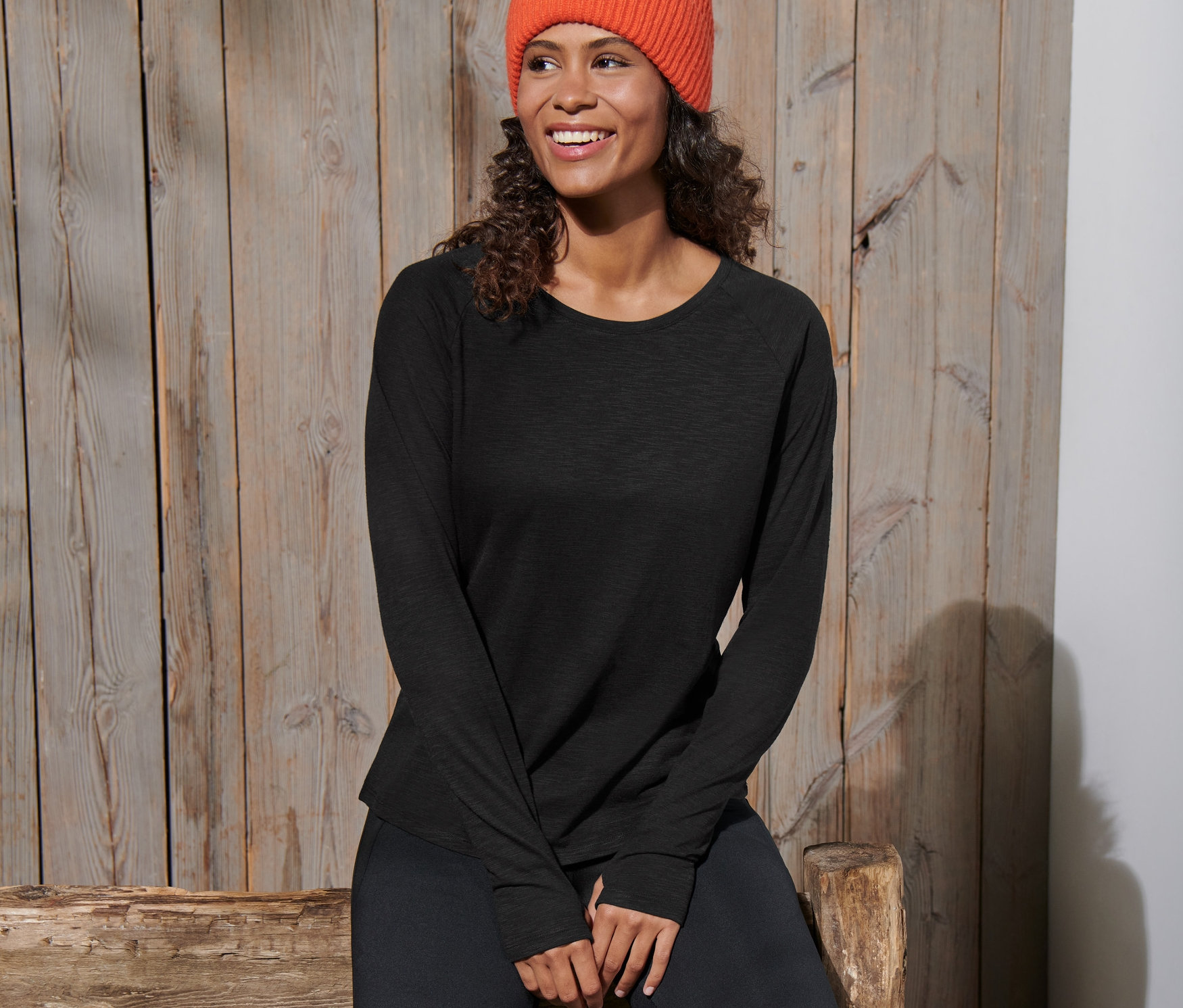 Kadın ahşap bir bankta oturuyor, üzerinde siyah uzun kollu spor tişörtü ve turuncu bir bere var.
