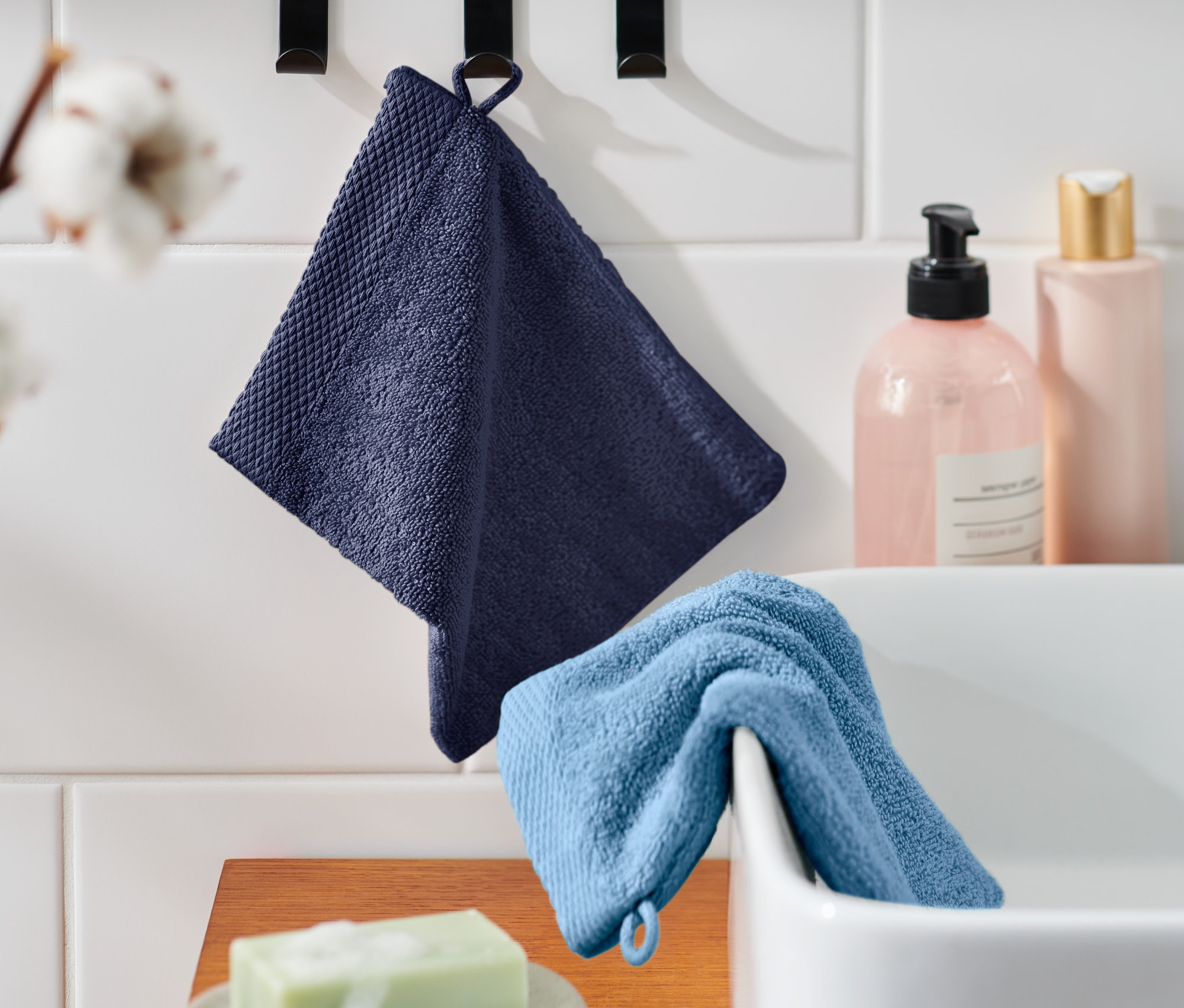 Lavabonun üzerinde açık mavi bir kesenin yanında kancada asılı duran koyu mavi bir kese. Arka planda kozmetik şişeleri ve çiçekler. 2 Adet Kese.