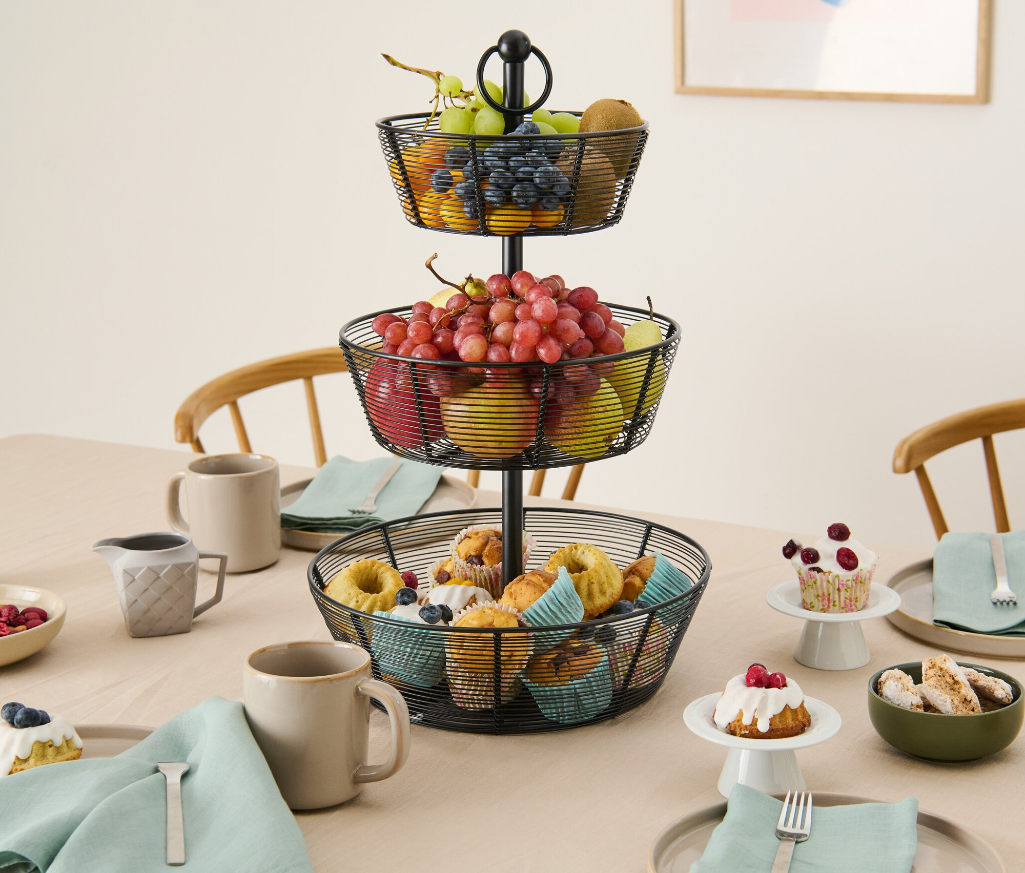 Masanın üzerinde meyve ve muffinlerle dolu üç katlı bir katlı sunum standı duruyor.