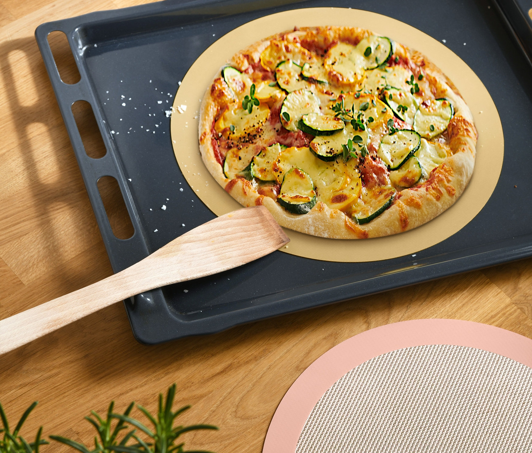 Fırın tepsisinde yuvarlak pişirme matı üzerinde pizza, yanında tahta spatula ve yanında ikinci bir yuvarlak pişirme matı.