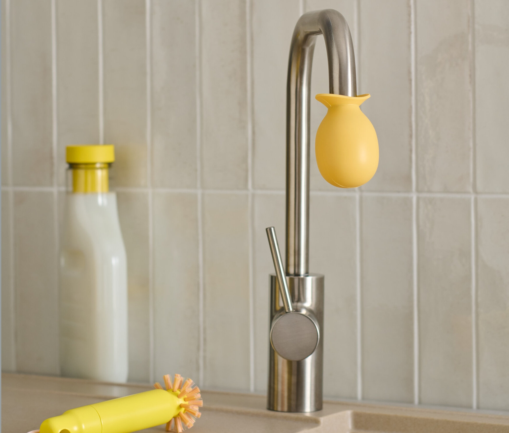 Paslanmaz çelik musluğun üzerinde sarı bir Musluk Kireç Çözme Başlığı var. Arka planda bir şişe ve sarı bir fırça bulunuyor.