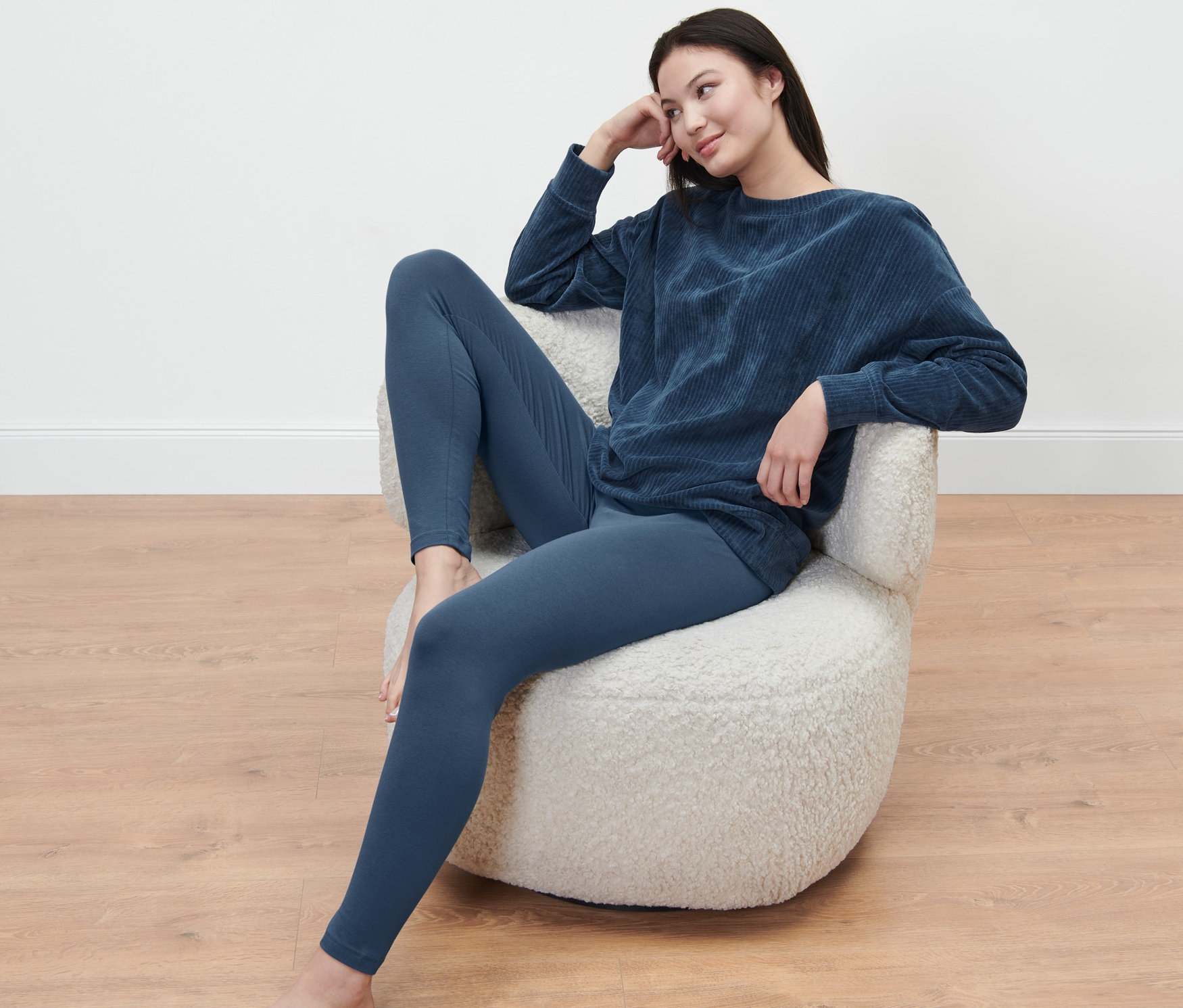 Kadın koltukta oturuyor, üzerinde taytlı pijama takımı var.