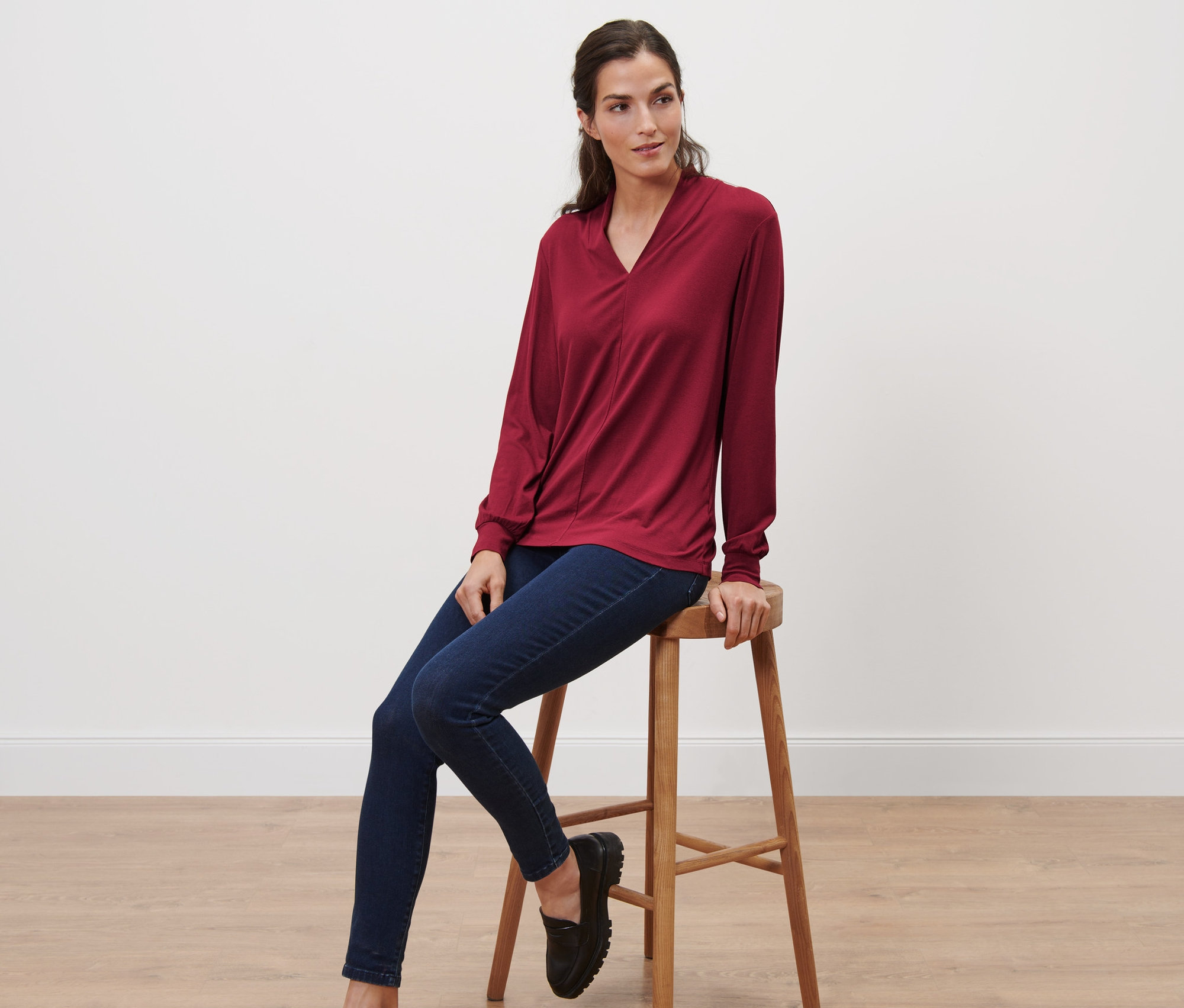 Kadın taburede oturuyor ve bordo, uzun kollu, V yaka bir üst giyiyor.