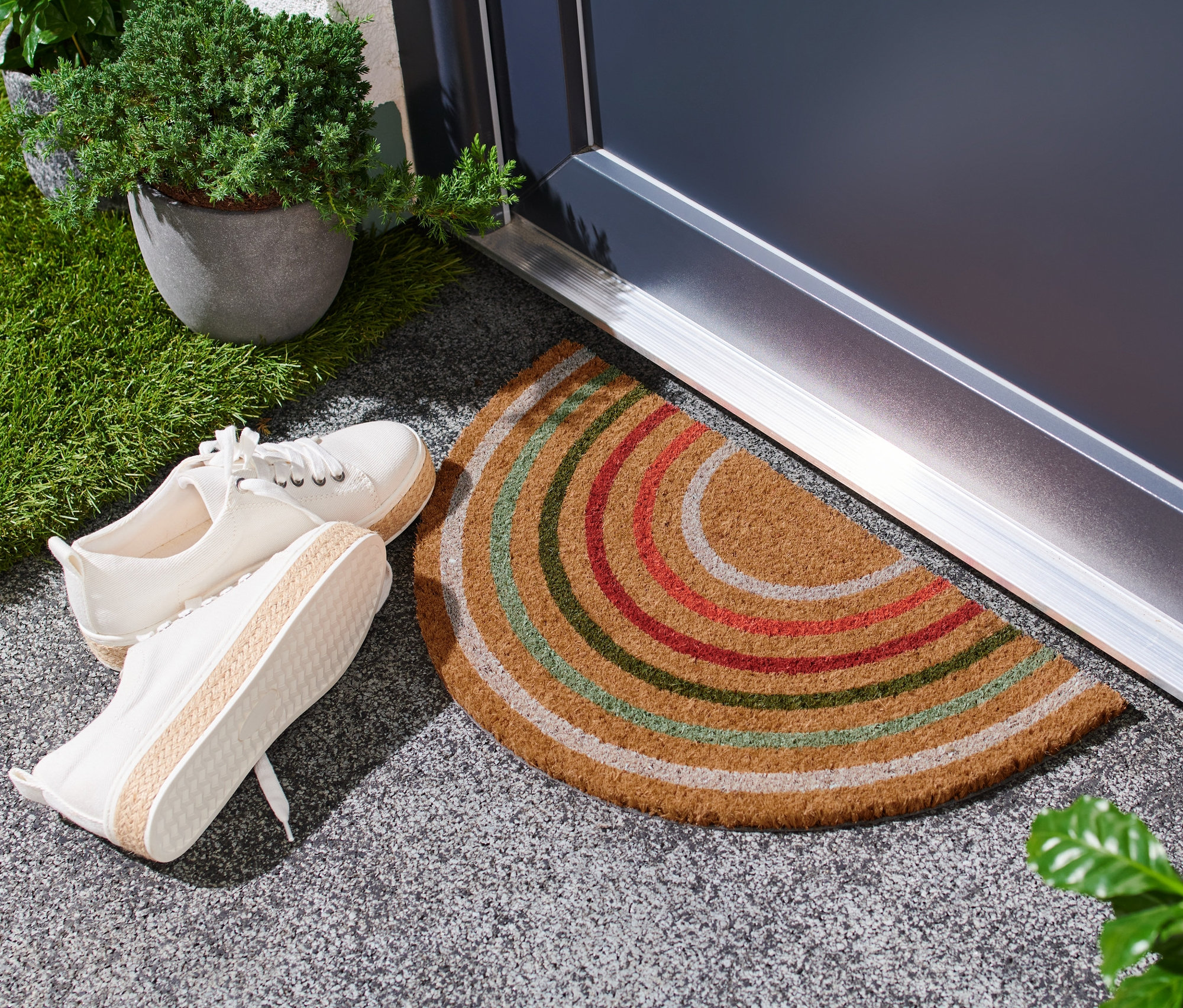 Bir kapının önünde renkli şeritli bir Hindistan cevizi paspas duruyor, yanında bir çift beyaz ayakkabı var.