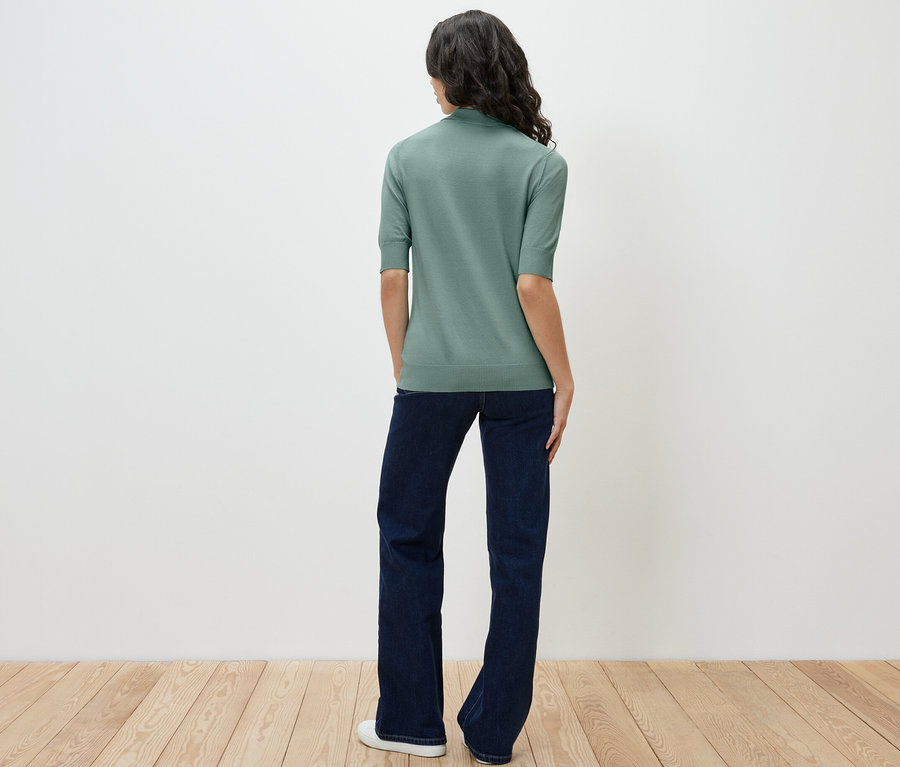 Bir kadın kameraya arkası dönük duruyor, açık yeşil ince triko polo ve kot pantolon giyiyor.