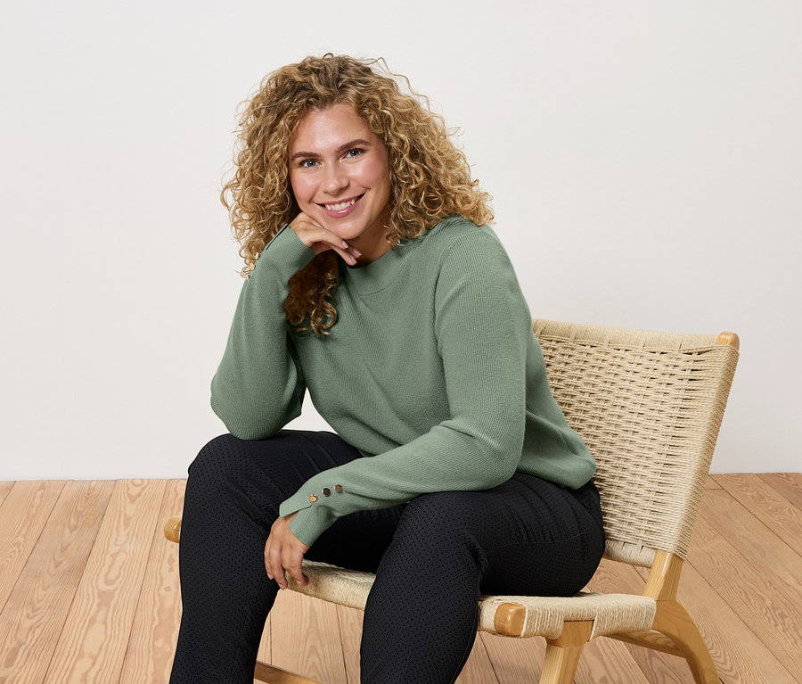 Kadın yeşil kazak ve siyah pantolonla sandalyede oturuyor.