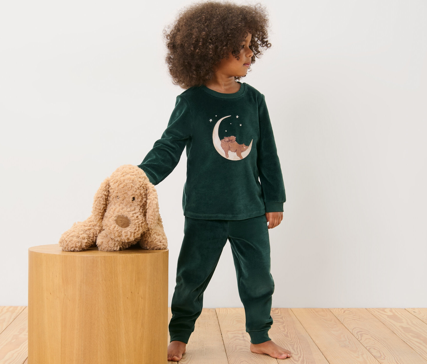 Çocuk, ahşap taburede peluş bir köpeğin yanında Niki pijamayla duruyor.