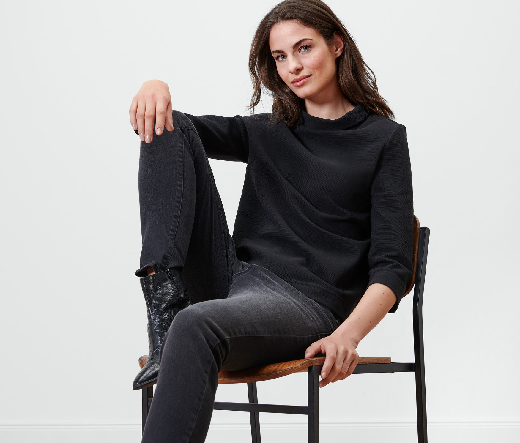 Kadın sandalyede oturuyor ve siyah, 3/4 kollu bir sweatshirt giyiyor.