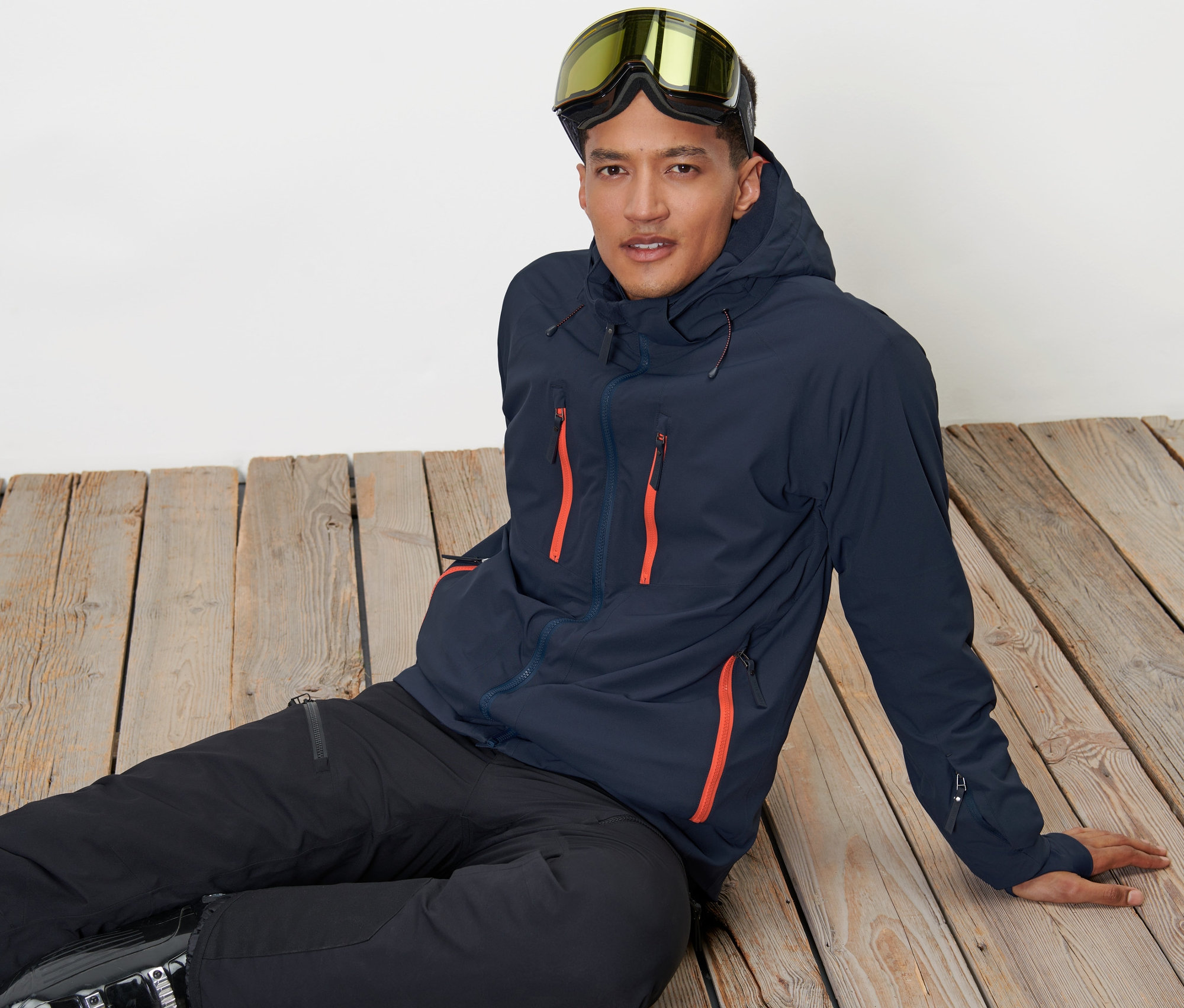 Adam ahşap zeminde oturuyor, Yüksek Teknoloji Kayak Montu ve kayak gözlüğü takıyor.