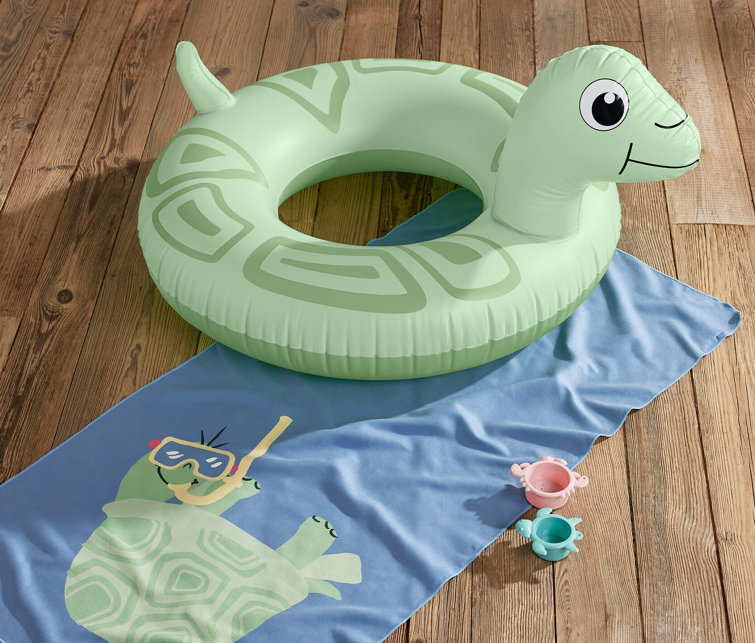 Ahşap zeminde kaplumbağa desenli mavi bir havlu ve iki oyuncak üzerinde yeşil bir »Kaplumbağa« deniz simidi duruyor.