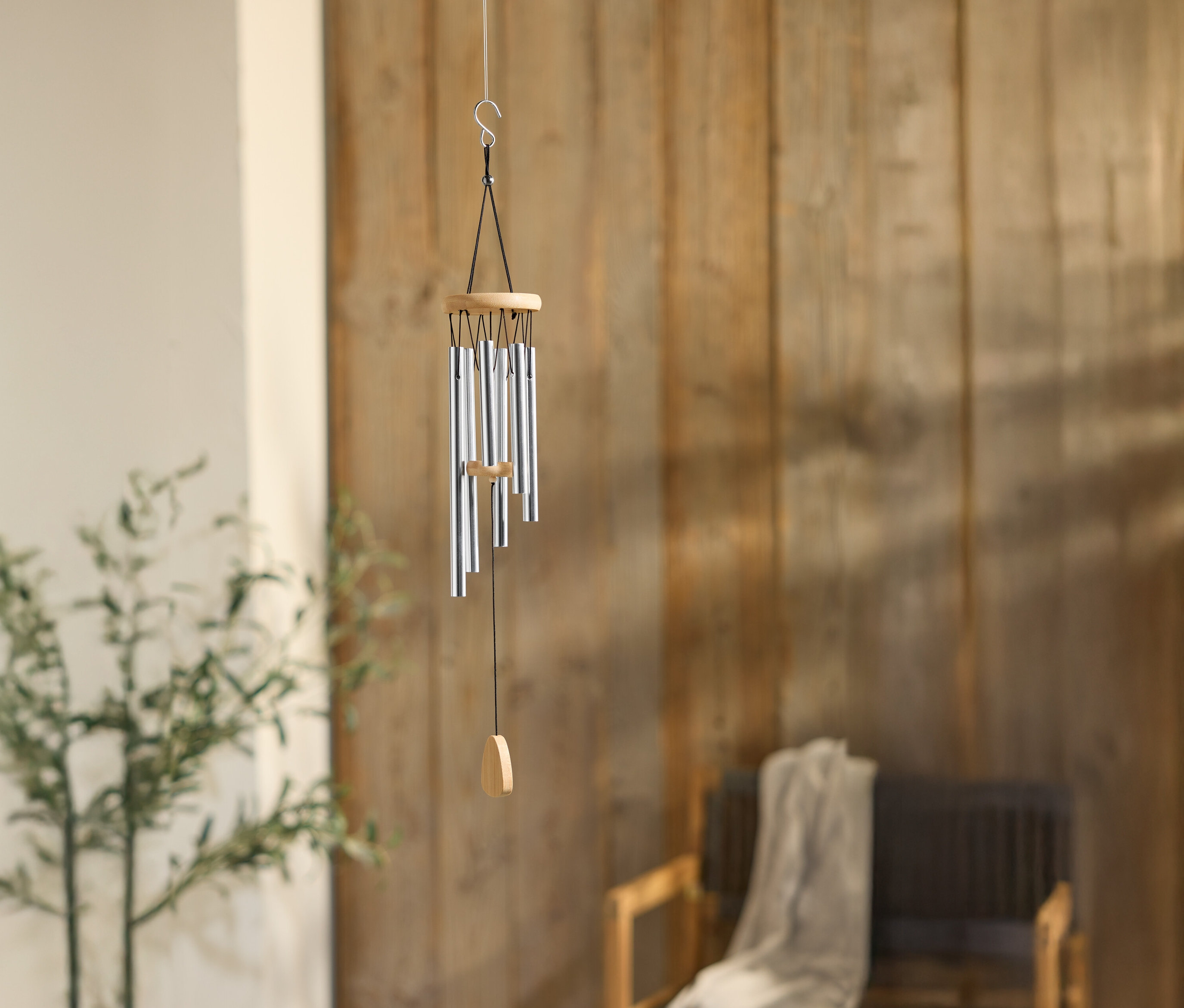 Gümüş borulu bir rüzgar çanı, ahşap bir duvarın önünde, bir bitki ve sandalyenin yanında asılı duruyor.