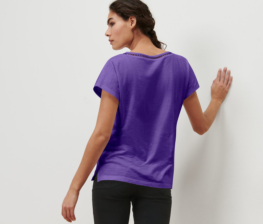 Örgülü saçlı bir kadın arkası dönük bir şekilde duvara yaslanmış, üzerinde mor modal karışımlı bir tişört var. 