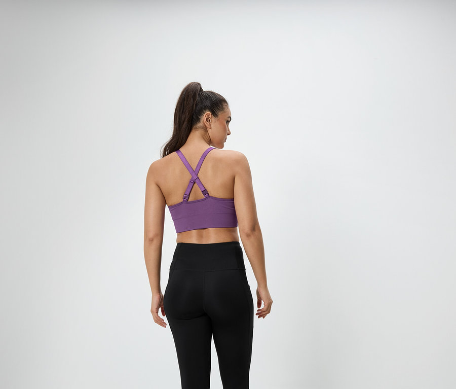 Kahverengi saçları at kuyruğu yapılmış bir kadın kameraya arkası dönük duruyor, mor bir spor sütyeni ve siyah tayt giyiyor.