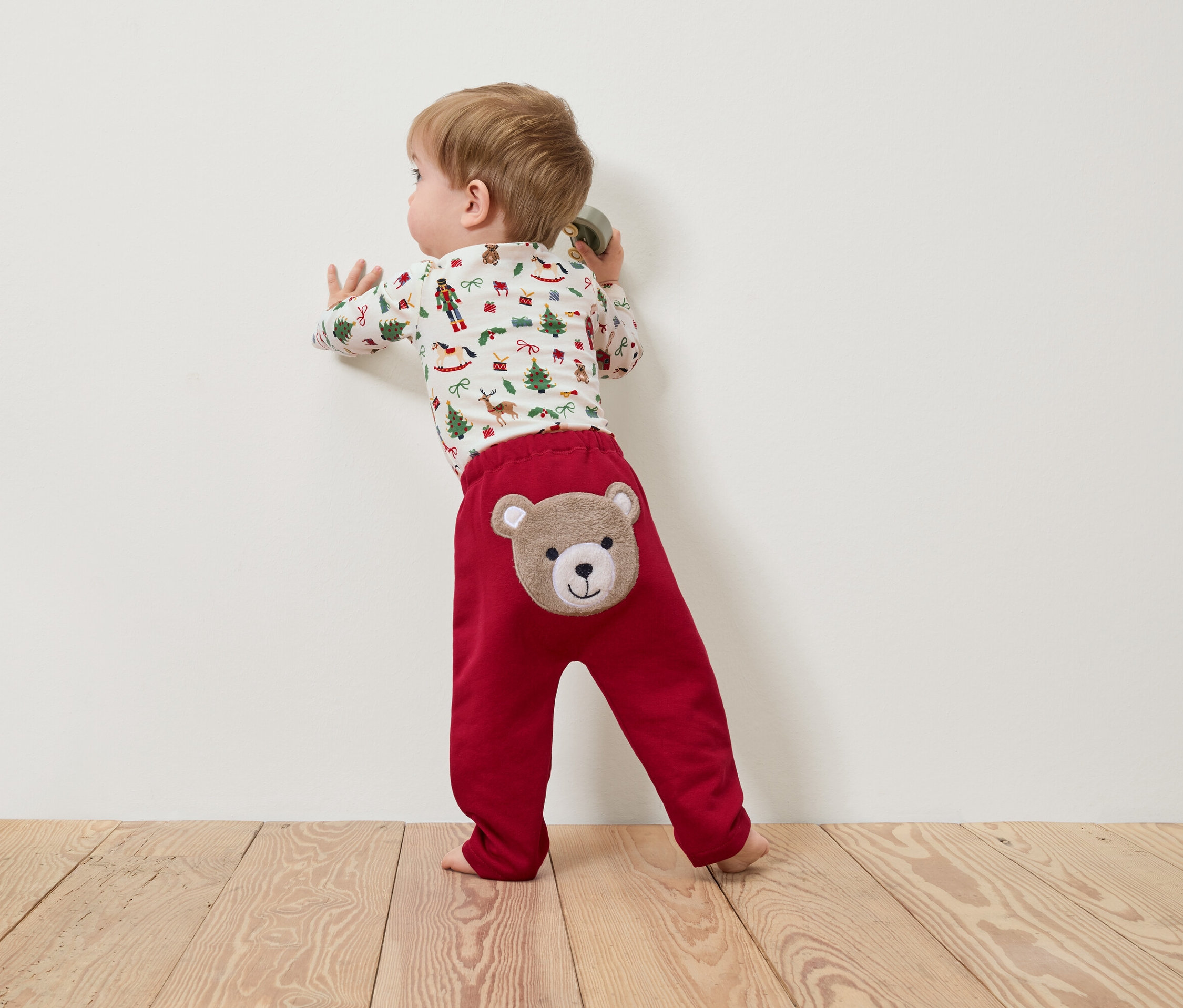 Bir çocuk duvara yaslanmış, üzerinde dikilmiş ayı kafası olan kırmızı bir pantolon ve Noel temalı bir tişört giyiyor.