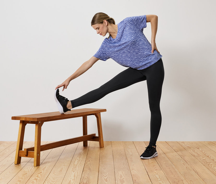 Mavi tişört ve siyah taytlı bir kadın, bacağını tahta bir bankın üzerine koyarak geriniyor.