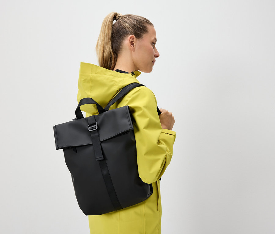 Sarışın saçları at kuyruğu yapılmış bir kadın sarı bir ceket ve siyah bir sırt çantası taşıyor.