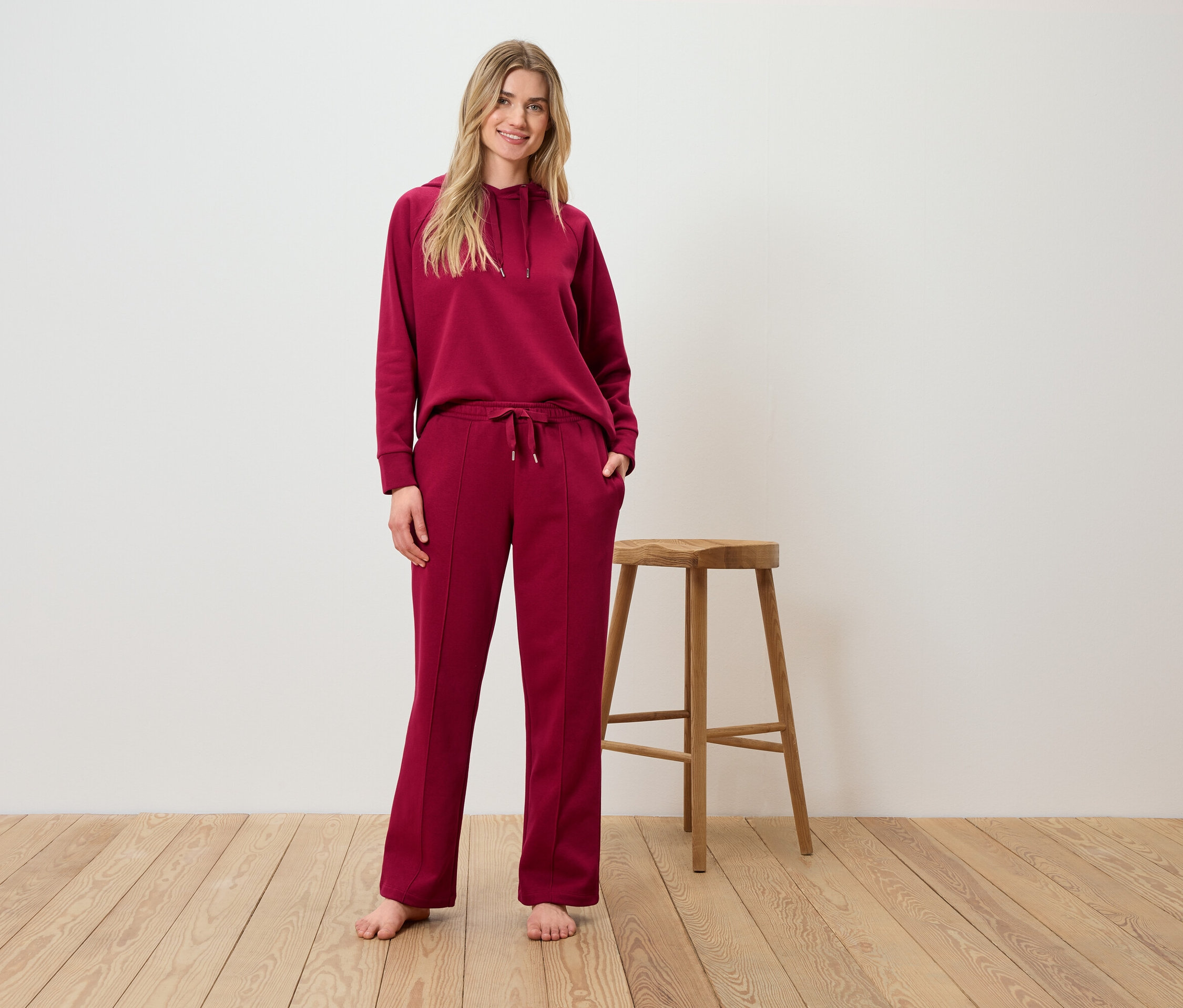 Bir kadın, bordo renkli kapüşonlu ve pantolonlu bir takım elbise içinde ahşap bir taburenin önünde poz veriyor.