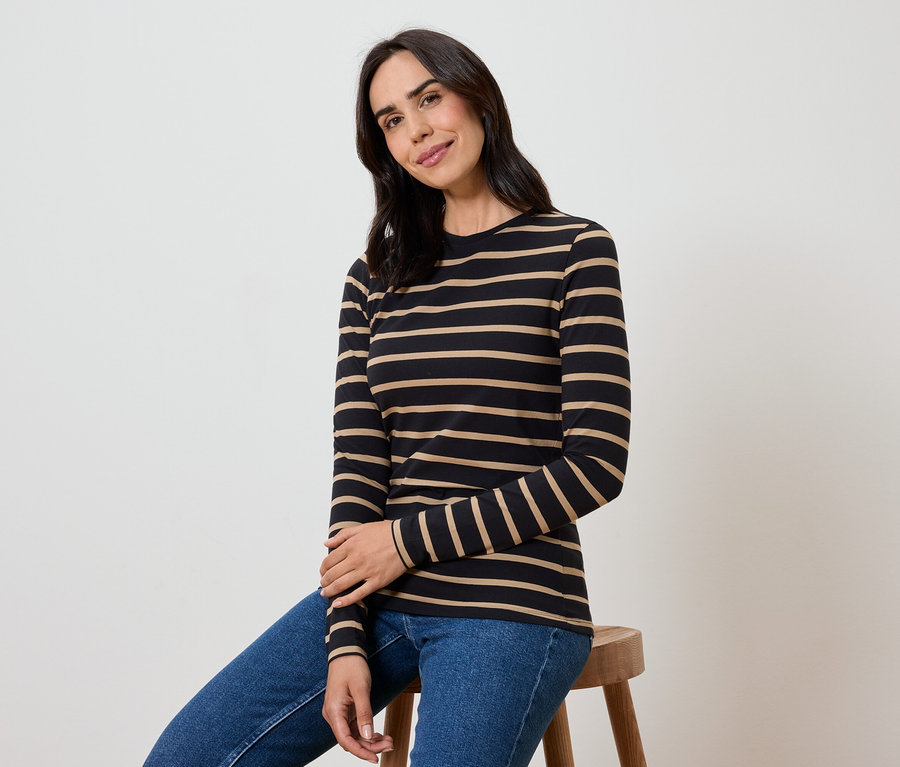 Bir kadın taburede oturuyor, siyah ve bej çizgili uzun kollu bir tişört ve mavi kot pantolon giyiyor.