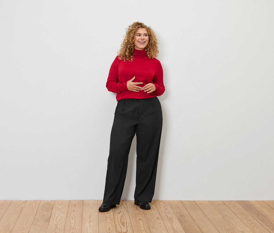 Kıvırcık saçlı bir kadın ayakta duruyor, kırmızı balıkçı yaka kazak ve siyah pantolon giyiyor.