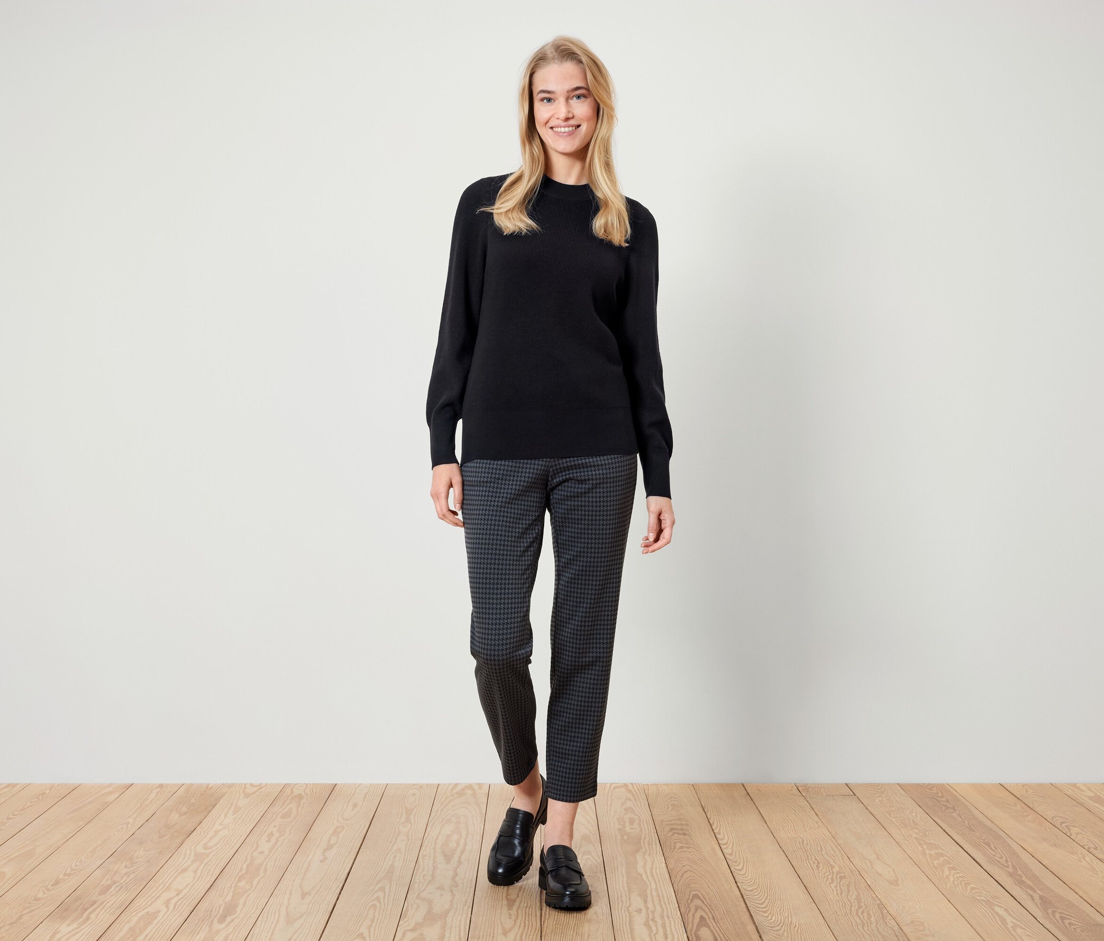 Bir kadın ayakta duruyor ve gülümsüyor, siyah bir kazak, desenli pantolon ve siyah loafer ayakkabılar giyiyor.