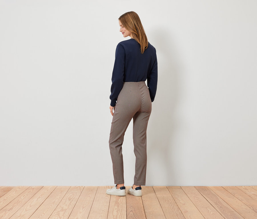 Kadın kameraya arkası dönük duruyor, lacivert bir kazak, kahverengi ekose pantolon ve beyaz spor ayakkabı giyiyor.