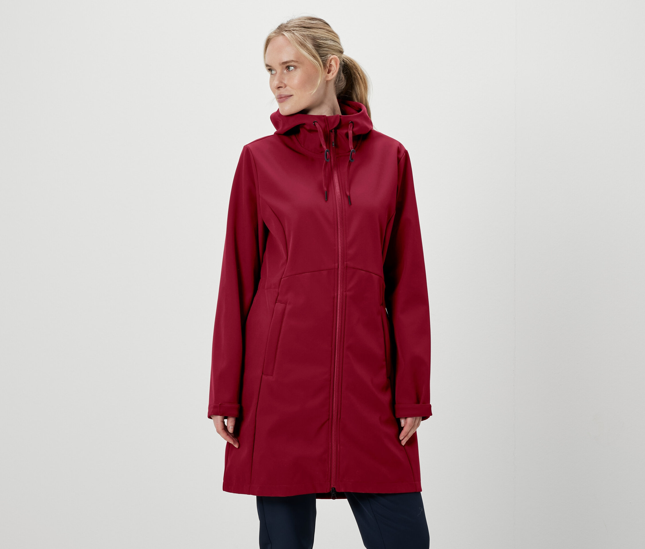 Beyaz arka plan önünde bordo renkli, kapüşonlu, fermuarlı bir ceket ve pantolon giyen sarışın kadın.