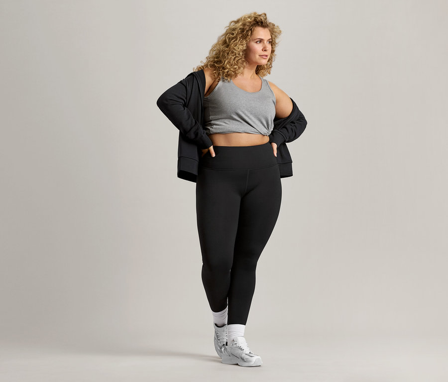 Kıvırcık saçlı kadın gri bir atlet, siyah bir ceket ve siyah tayt giyiyor.