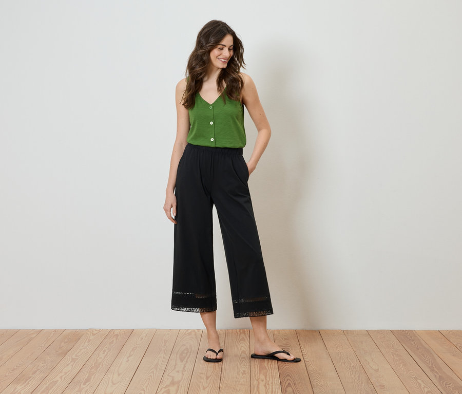 Kadın elleri cebinde duruyor, yeşil bir bluz ve etek ucunda dantel detaylı siyah bir culottes pantolon giyiyor.