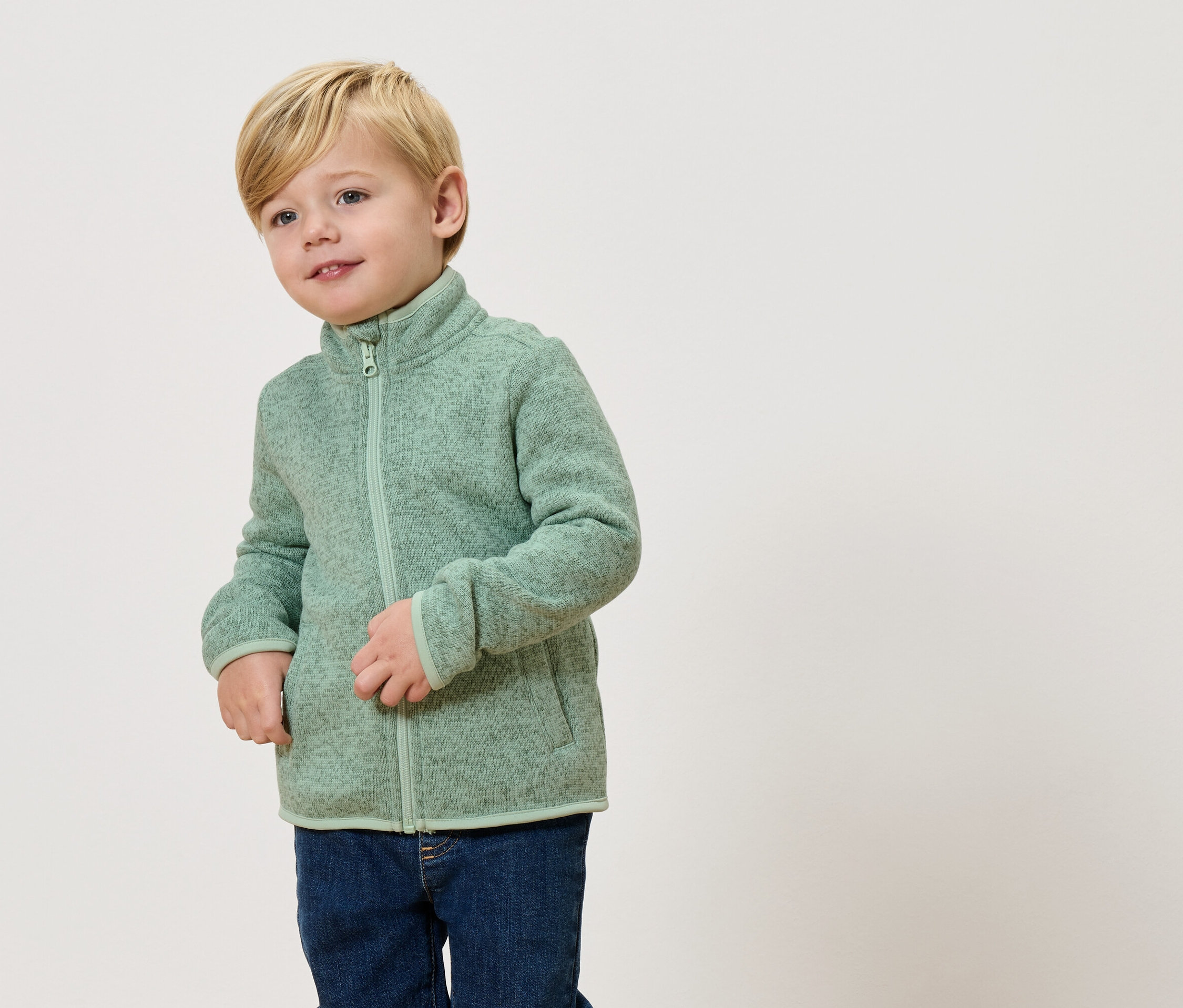 Sarışın bir çocuk açık yeşil bir polar ceket ve kot pantolon giyiyor.