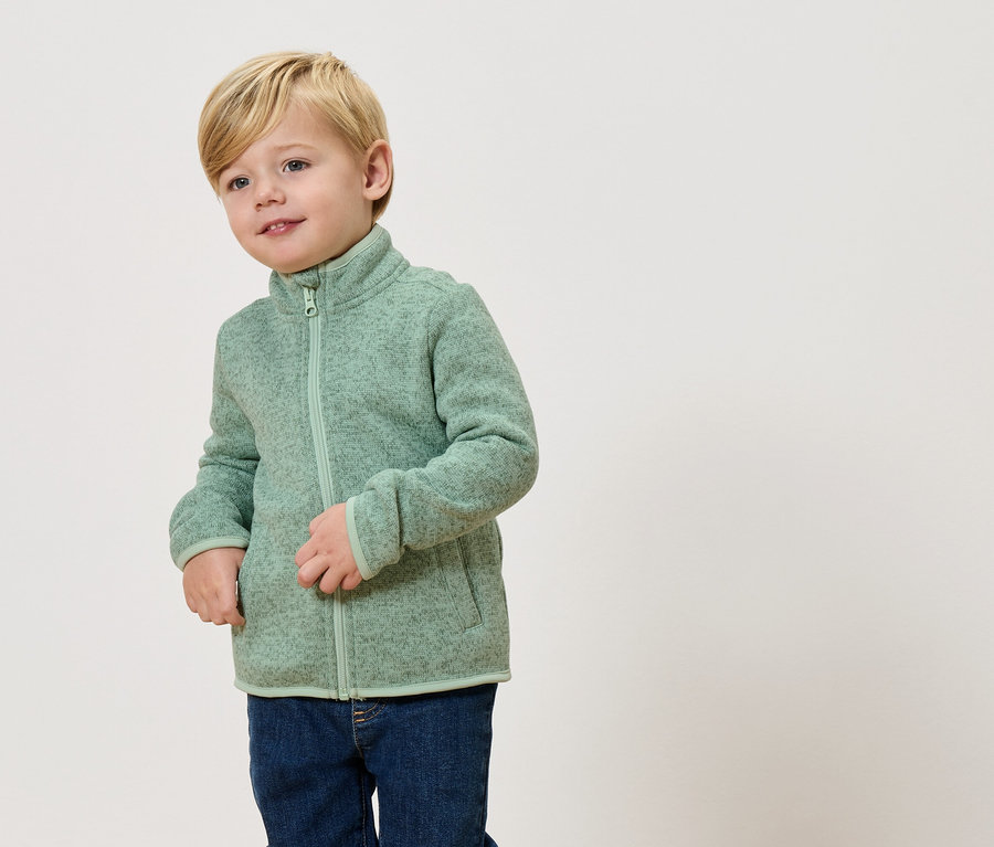 Sarışın bir çocuk açık yeşil bir polar ceket ve kot pantolon giyiyor.