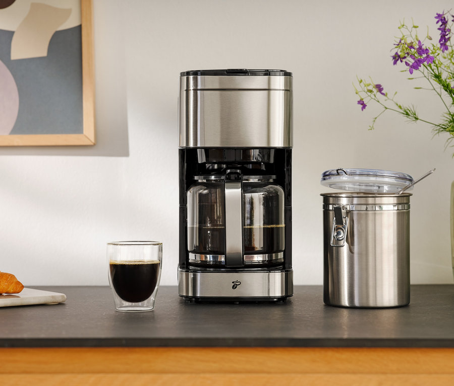 Kahve makinesi, bir bardak kahve, kruvasan ve saklama kabının yanında.