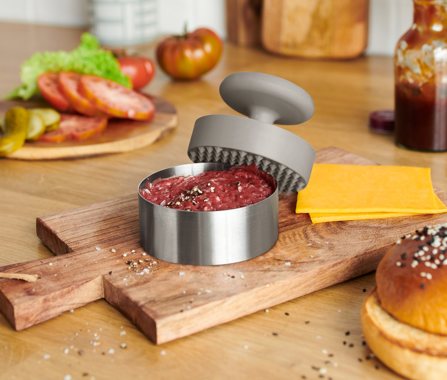Ahşap bir kesme tahtası üzerinde bir hamburger ekmeği, peynir ve içinde et bulunan açık bir burger presi duruyor.