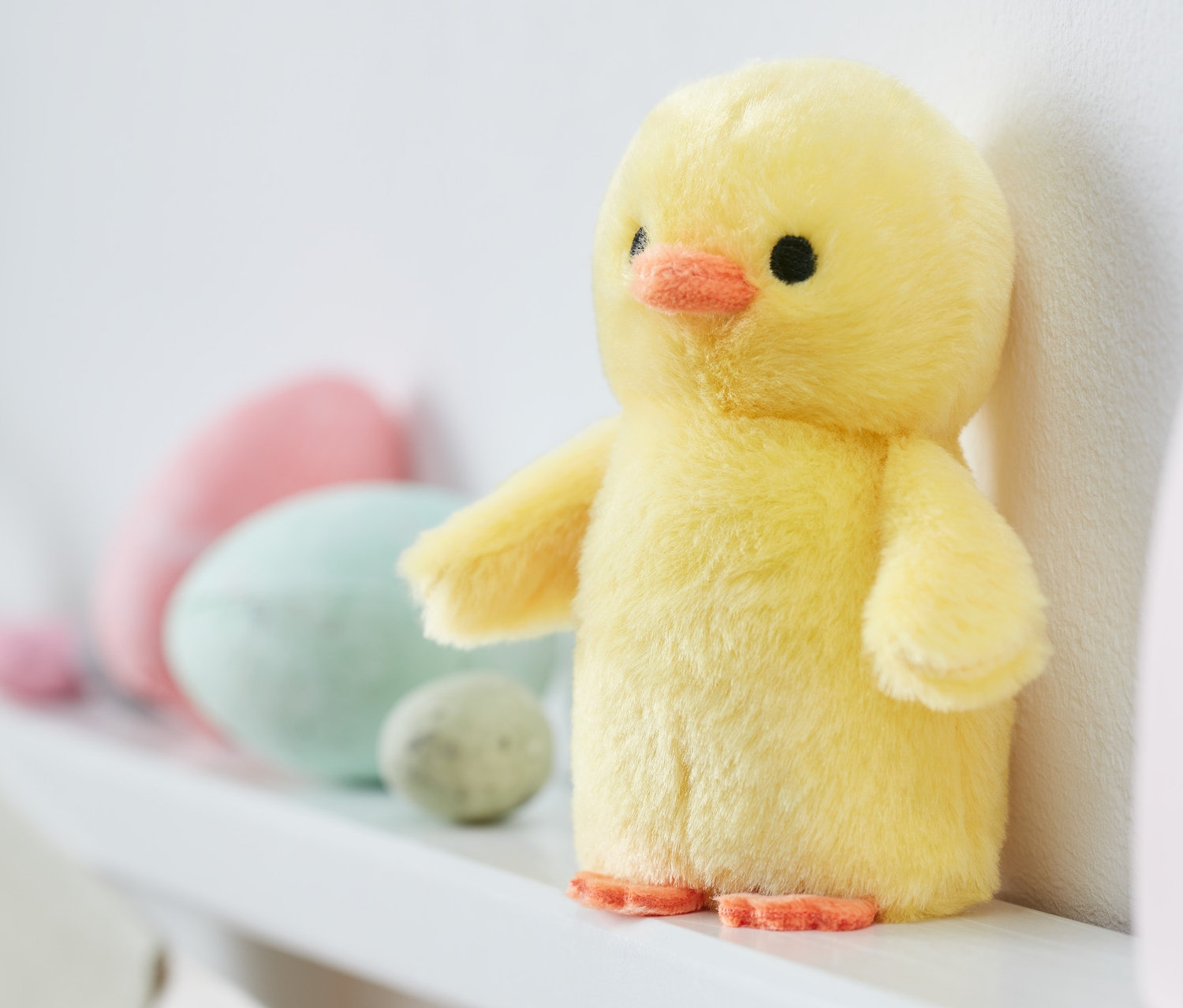 Sesli Civciv, dekoratif Paskalya yumurtalarının ve ahşap gökkuşağı oyuncağının yanında beyaz bir rafta duruyor.