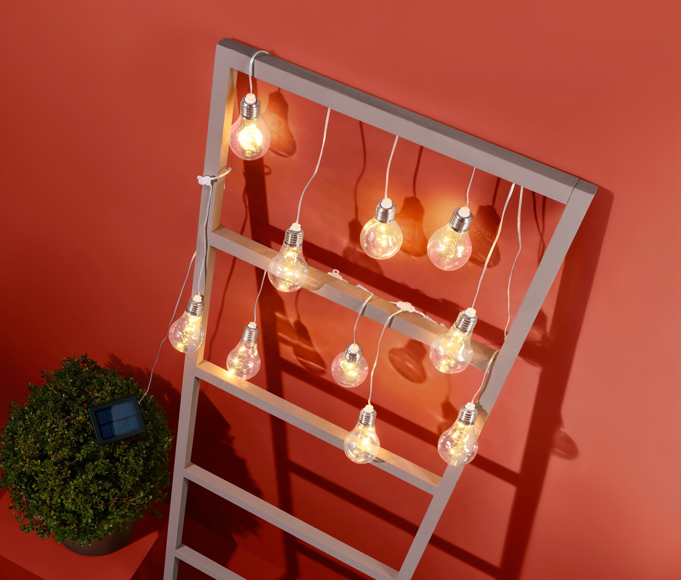 Güneş enerjili ışık zinciri, turuncu bir arka plan üzerinde küçük bir çalı yanında gri bir çerçeveye asılmıştır.