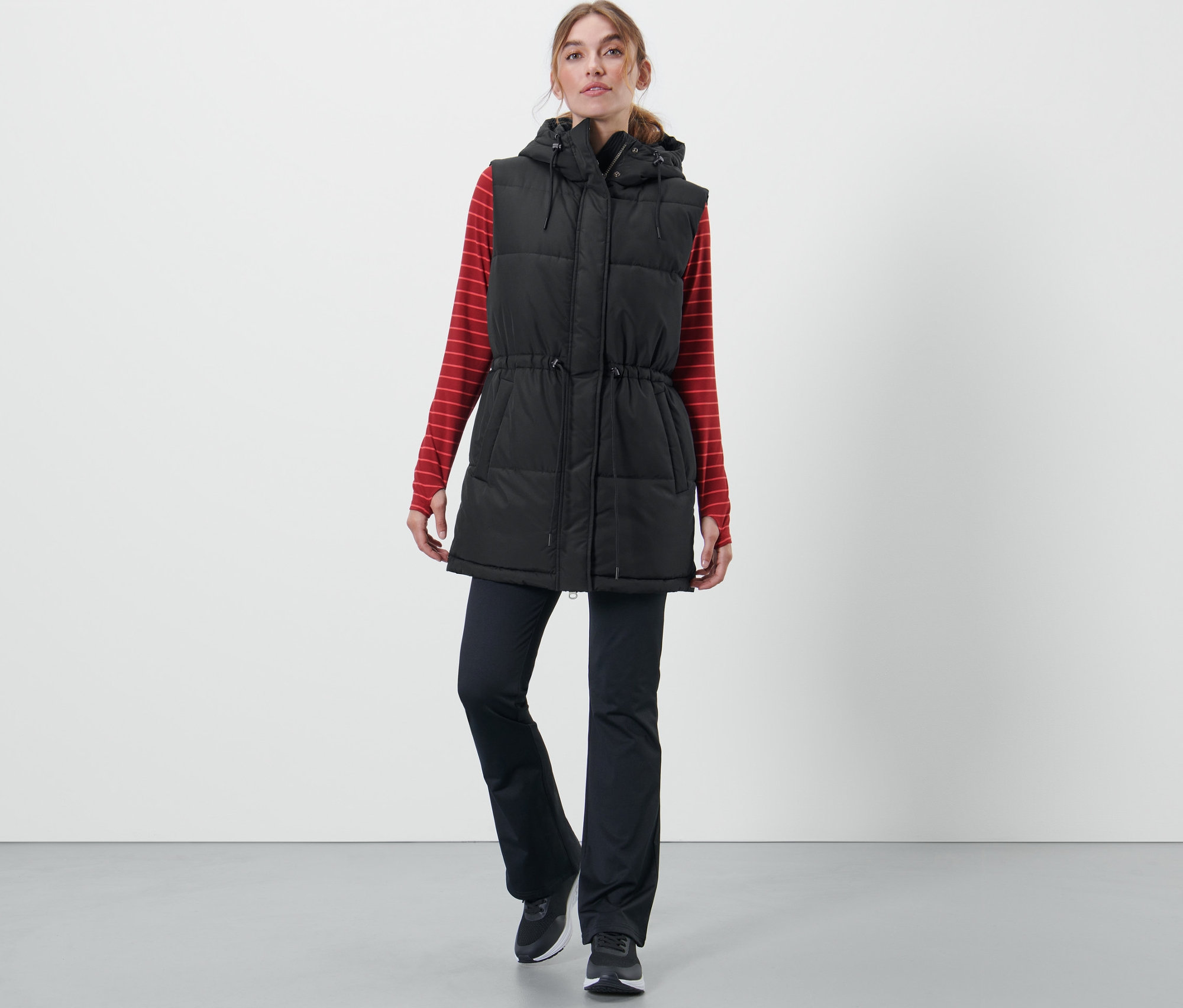 Kadın kapüşonlu kapitone yelek, kırmızı çizgili uzun kollu tişört ve siyah pantolon giyiyor.