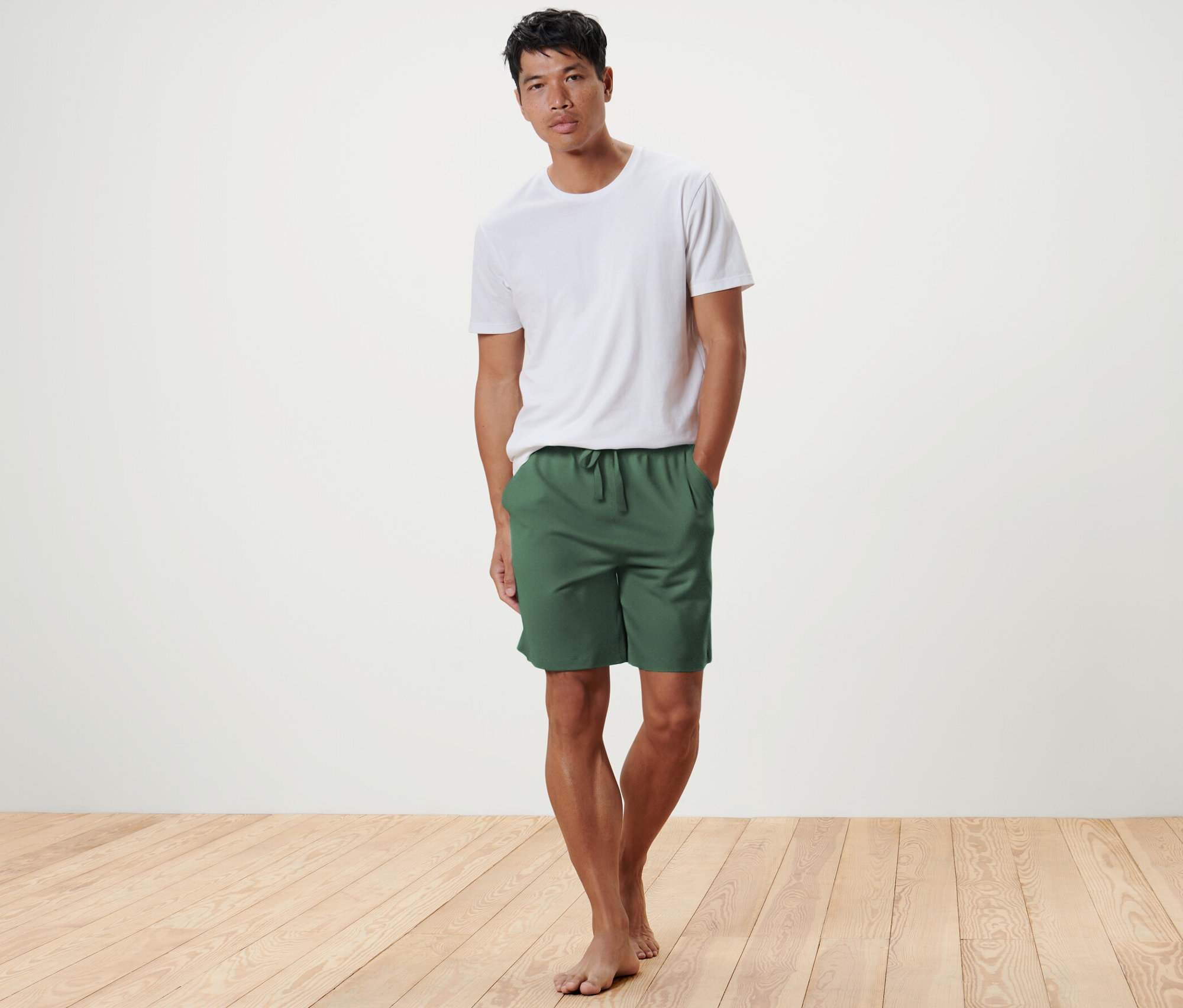 Ahşap zeminde zeytin yeşili Rahat Şort ve beyaz tişört ile poz veren adam.