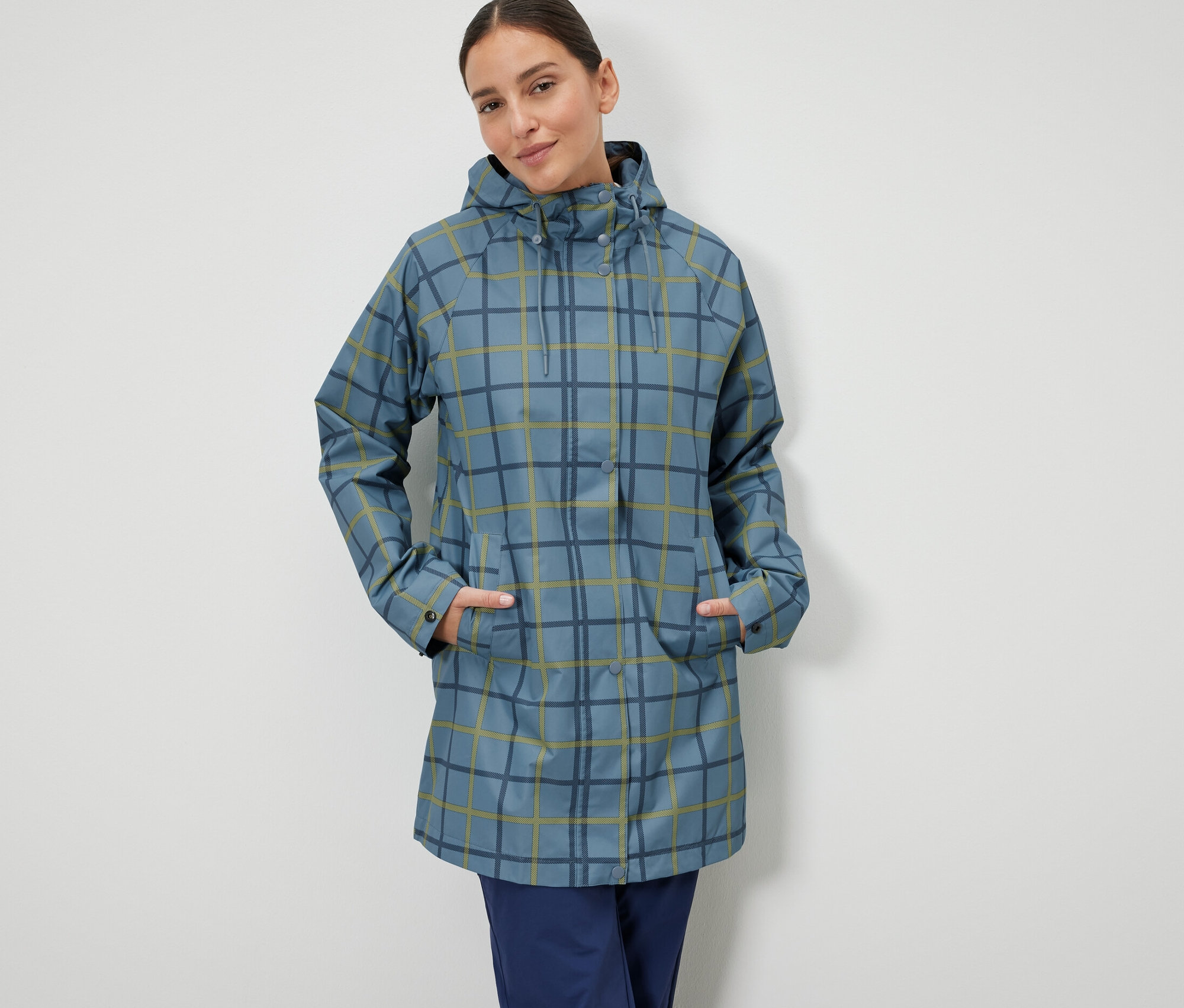Bir kadın, elleri mavi, kareli yağmurluğunun cebinde duruyor.