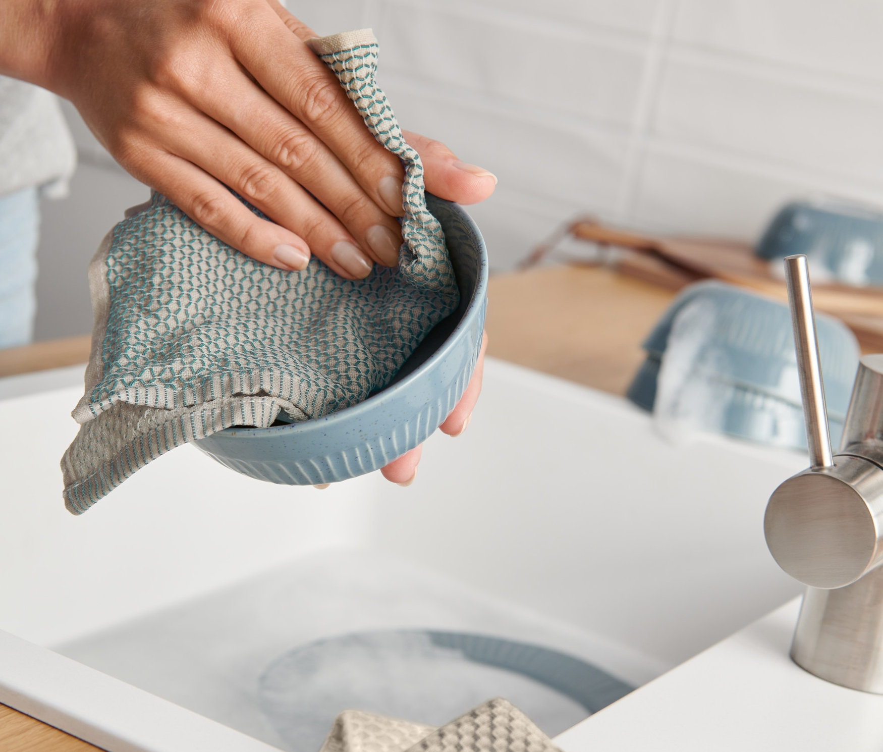 Bir kadın mavi bir bulaşık beziyle mavi bir kaseyi yıkıyor, tezgahta katlanmış 3 Adet Organik Pamuklu Bulaşık Bezi duruyor.