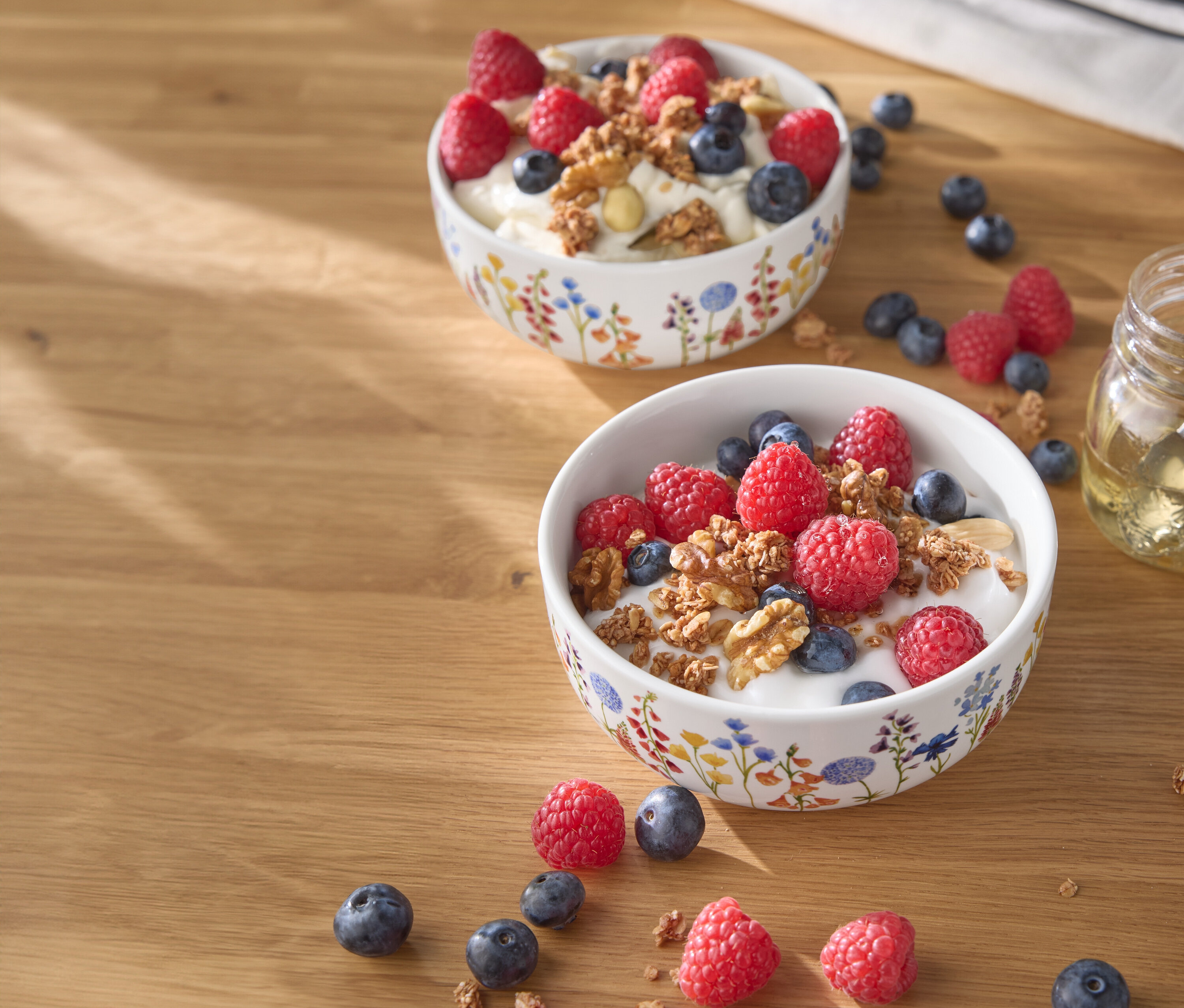 Ahşap bir masanın üzerinde ahududu, yaban mersini ve fındık bulunan iki kase yoğurt.