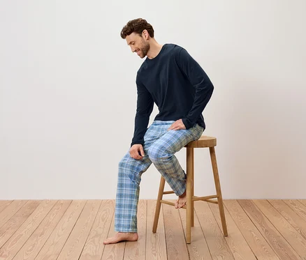 Adam ahşap bir taburede oturuyor, lacivert bir pijama ve mavi ekose pantolon giyiyor.