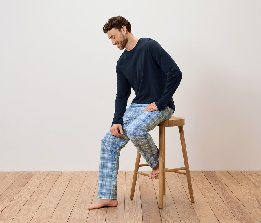 Adam ahşap bir taburede oturuyor, lacivert bir pijama ve mavi ekose pantolon giyiyor.