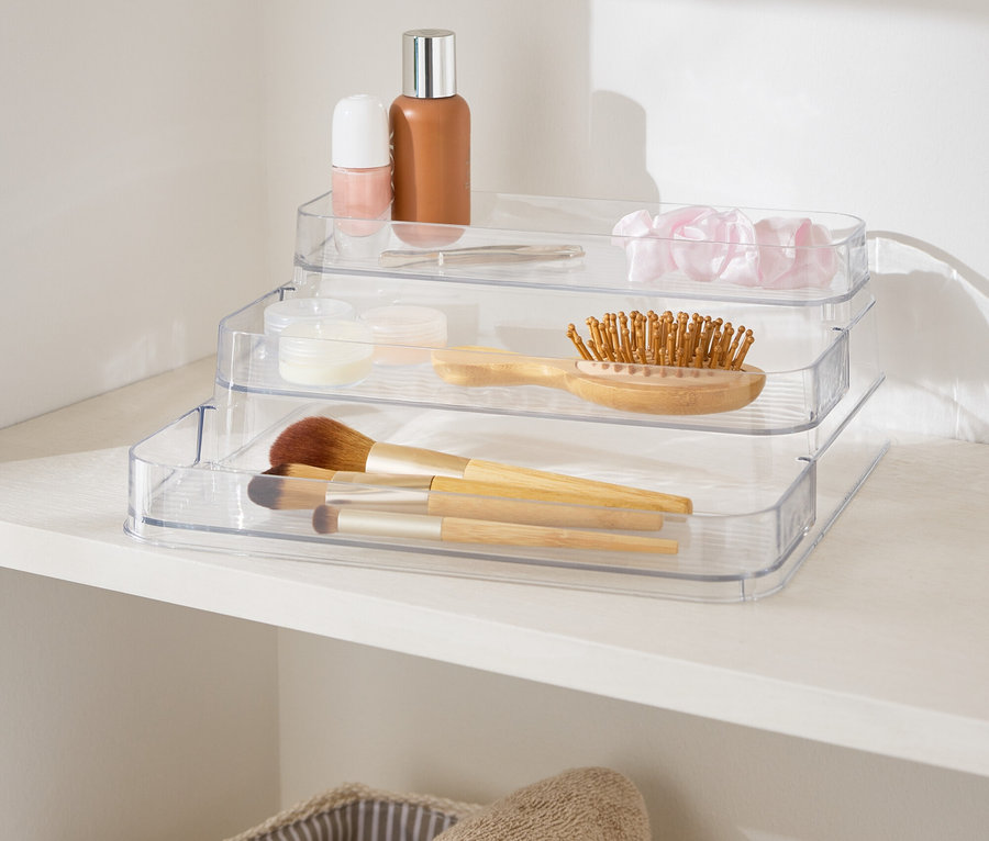Beyaz bir rafta, havlu sepetlerinin yanında duran üç katlı, şeffaf makyaj düzenleyici.