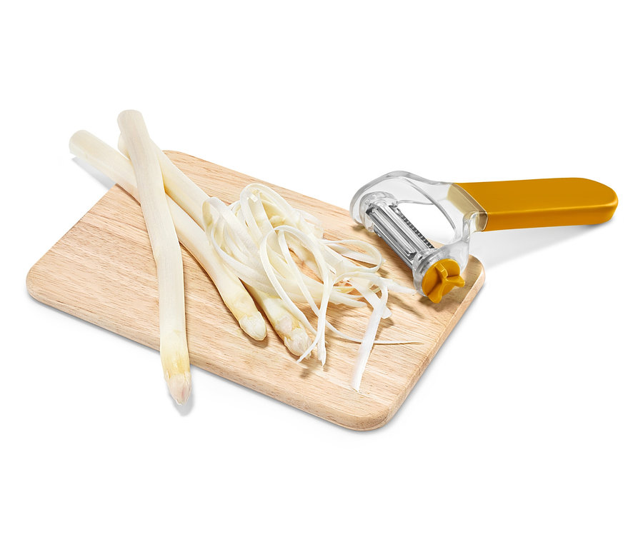 Ahşap bir kesme tahtası üzerinde üç adet beyaz kuşkonmaz sapı, kuşkonmaz talaşı ve sarı-gümüş bir sebze soyacağı bulunmaktadır.