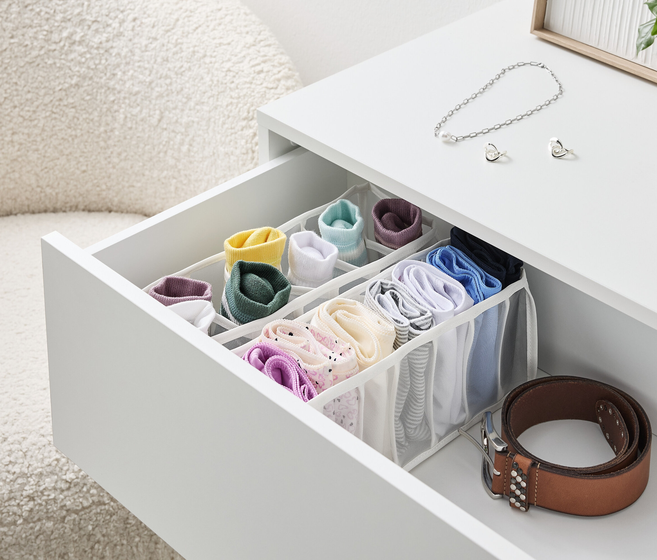 Beyaz bir masanın açık çekmecesi, içinde renkli çoraplar ve iç çamaşırları bulunan bir düzenleyiciyle dolu, yanında kahverengi bir kemer duruyor.