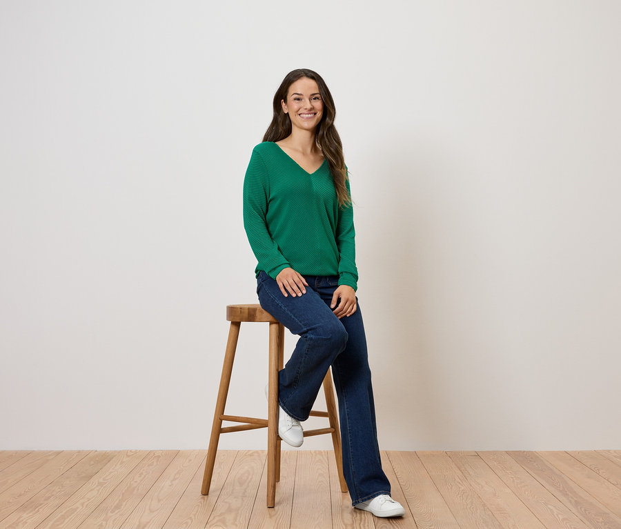 Bir kadın ahşap bir taburede oturuyor, üzerinde yeşil bir kazak ve kot pantolon var.