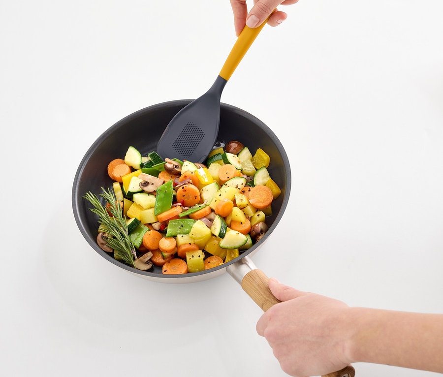 Eller, sarı, delikli bir spatula ile tavada sebzeleri karıştırıyor.