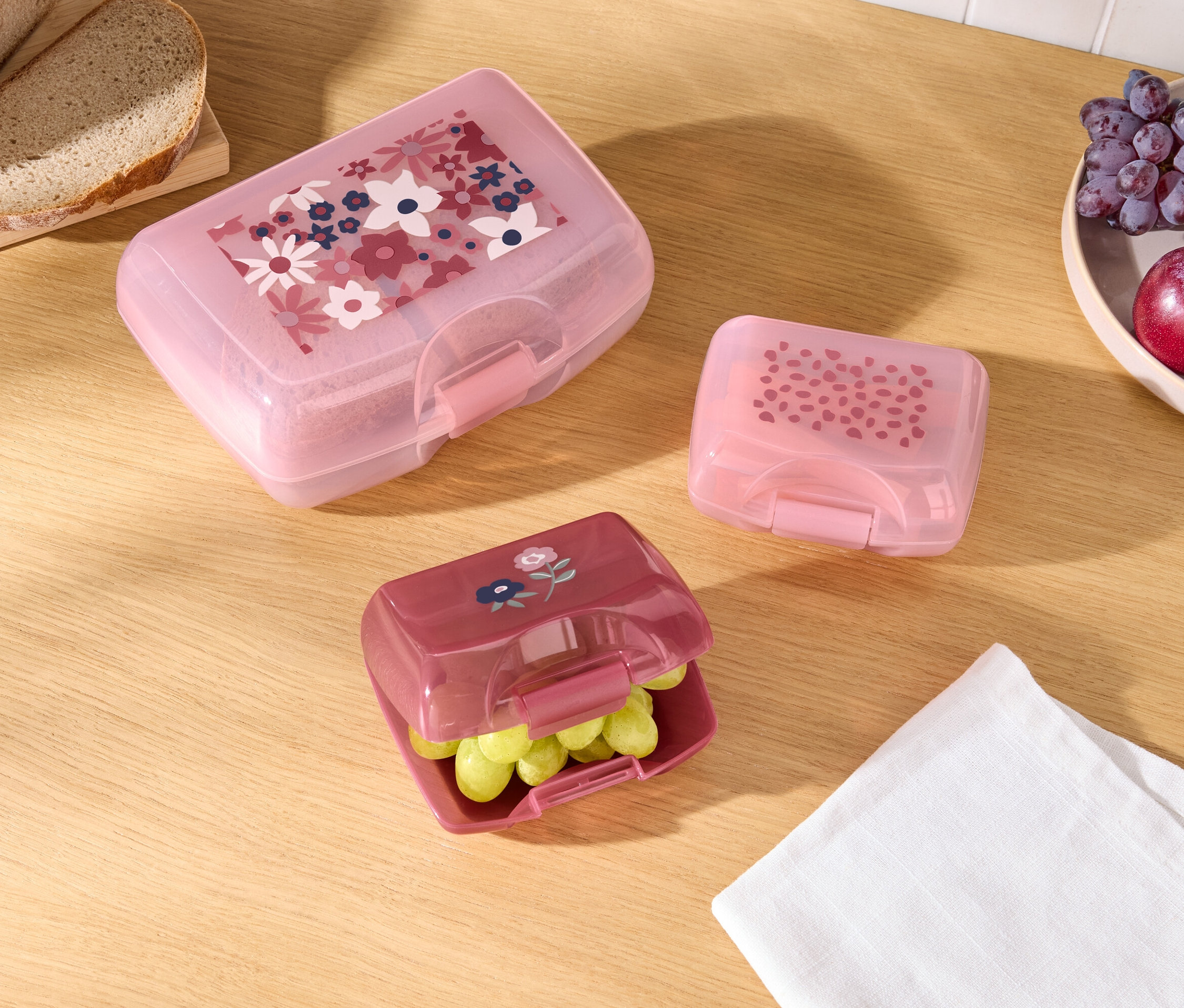 Ahşap bir masa üzerinde farklı desenlere sahip üç renkli yemek kutusu, ekmek ve meyve bulunmaktadır.