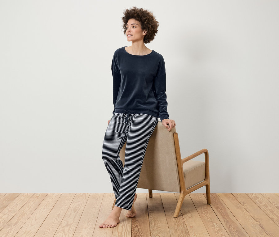 Afro saçlı bir kadın, ahşap zeminde bej bir koltuğa yaslanarak lacivert kadife bir üst ve çizgili bir pantolonla poz veriyor.